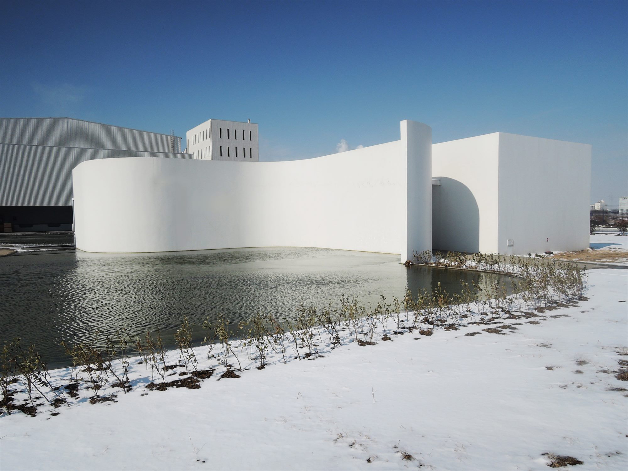 Curved white wall forming a welcoming gesture at the entrance of the salt laboratory, beside a frozen pond in winter snow